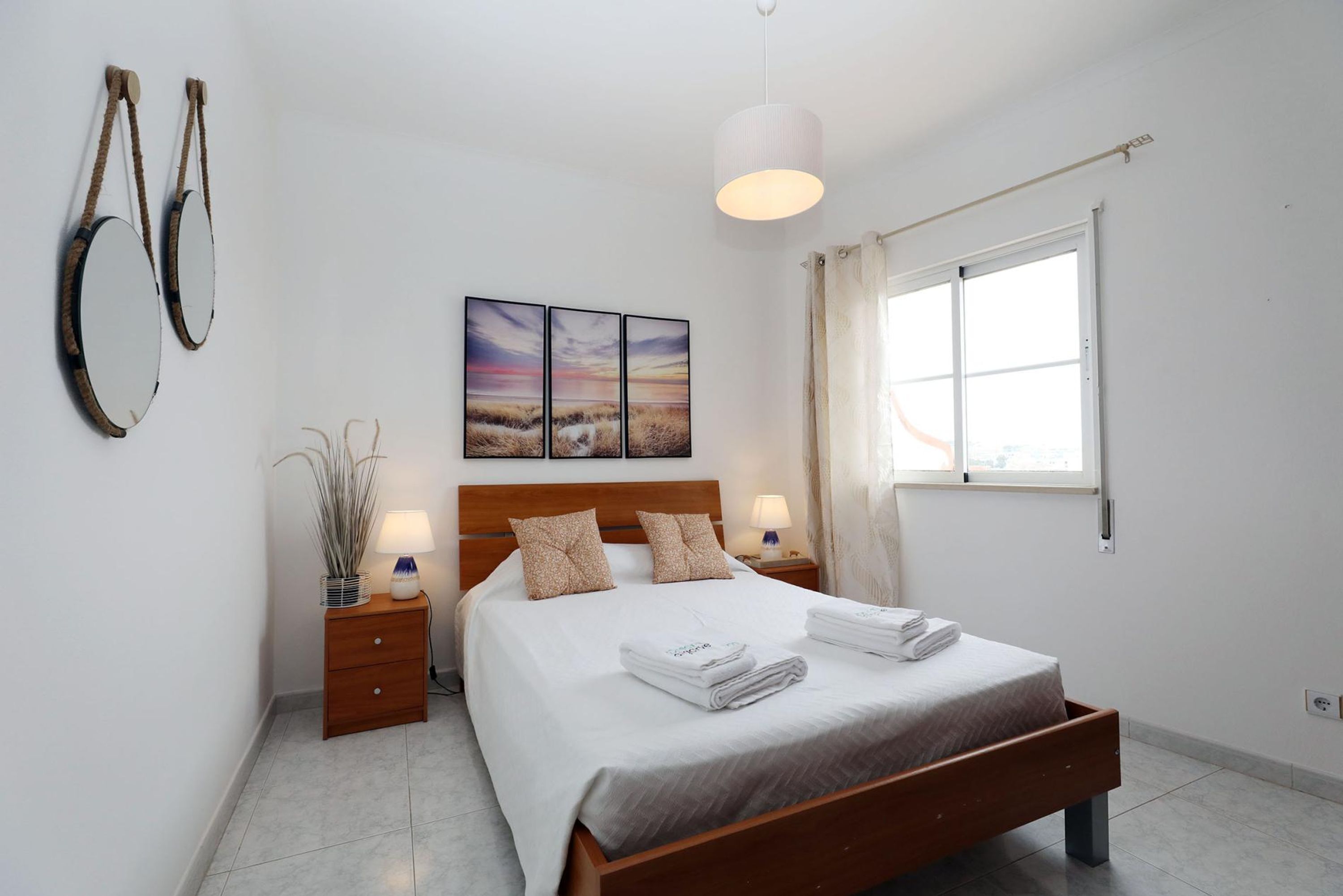 Une chambre à coucher avec un lit en bois, deux tables de chevet avec des lampes, une grande fenêtre et un miroir fixé au mur.