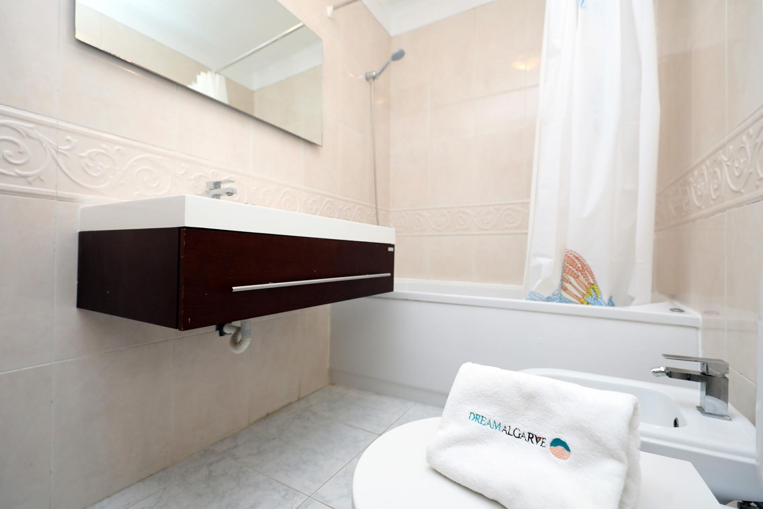 Un baño con lavamanos blanco, mueble de madera oscura y una toalla blanca con la inscripción "DREAMALGARVE".