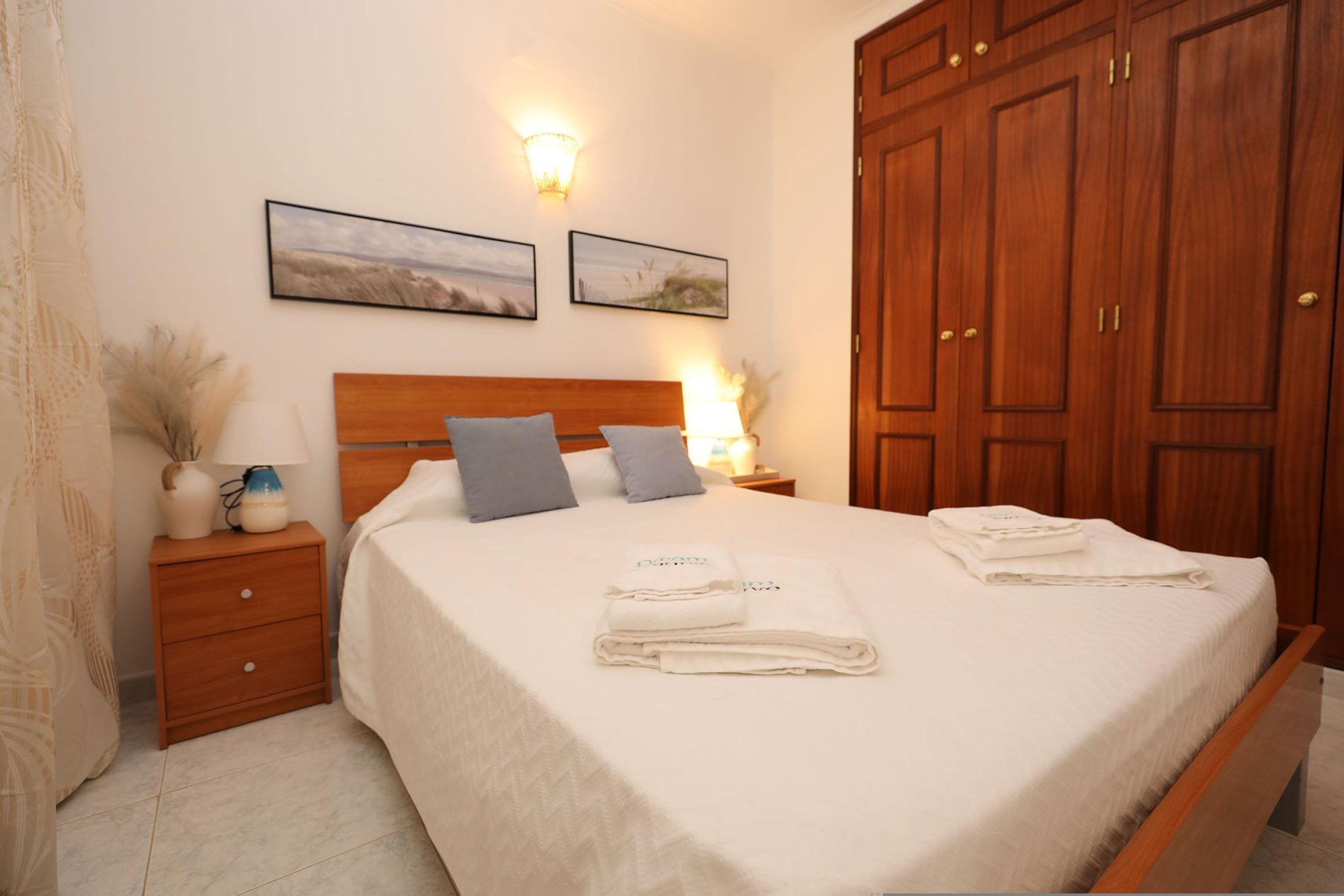 A neatly made bed with folded towels, a wooden nightstand with a lamp, and two framed pictures on the wall.