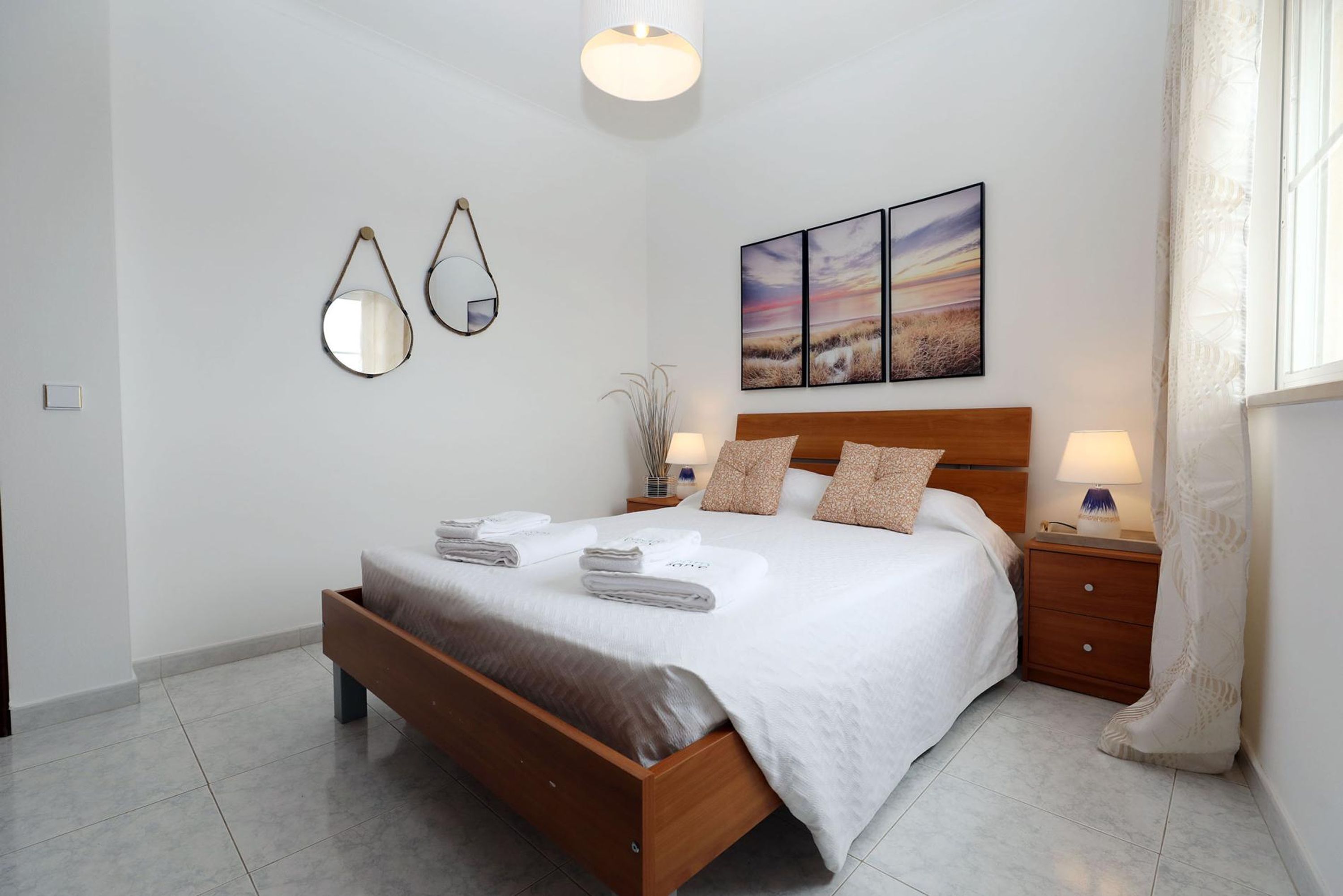Una cama bien tendida con un cabecero de madera, dos mesitas de noche con lámparas y un espejo fijado a la pared.