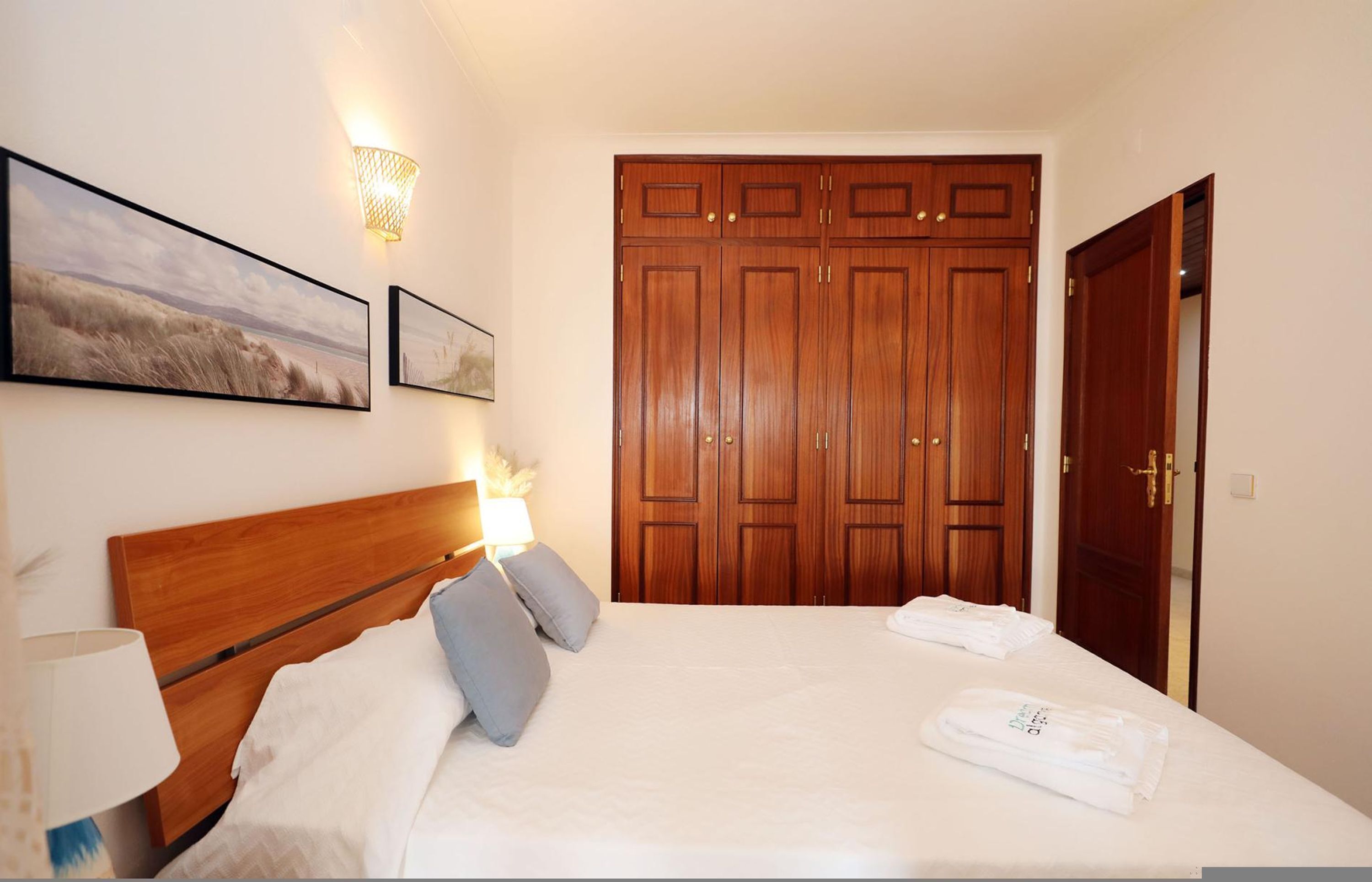 A hotel room with a wooden bed, a nightstand, a wall-mounted lamp, and a large wardrobe.