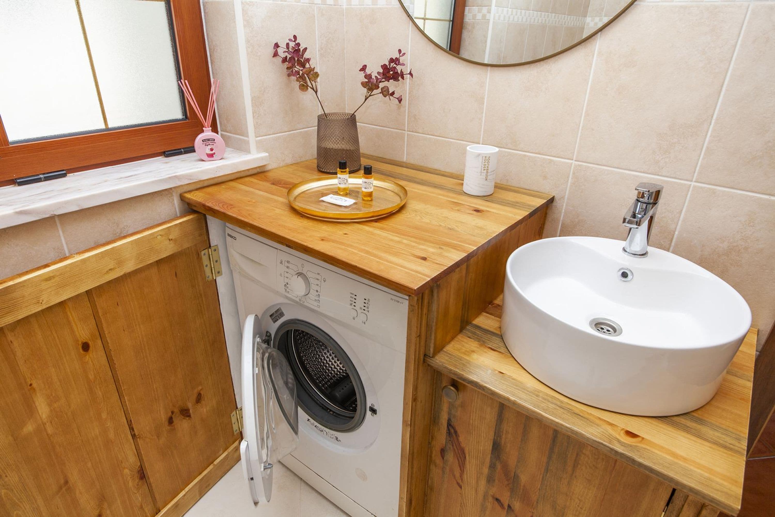 A compact bathroom with a wooden vanity, a sink, a washing machine, and a window with a view.