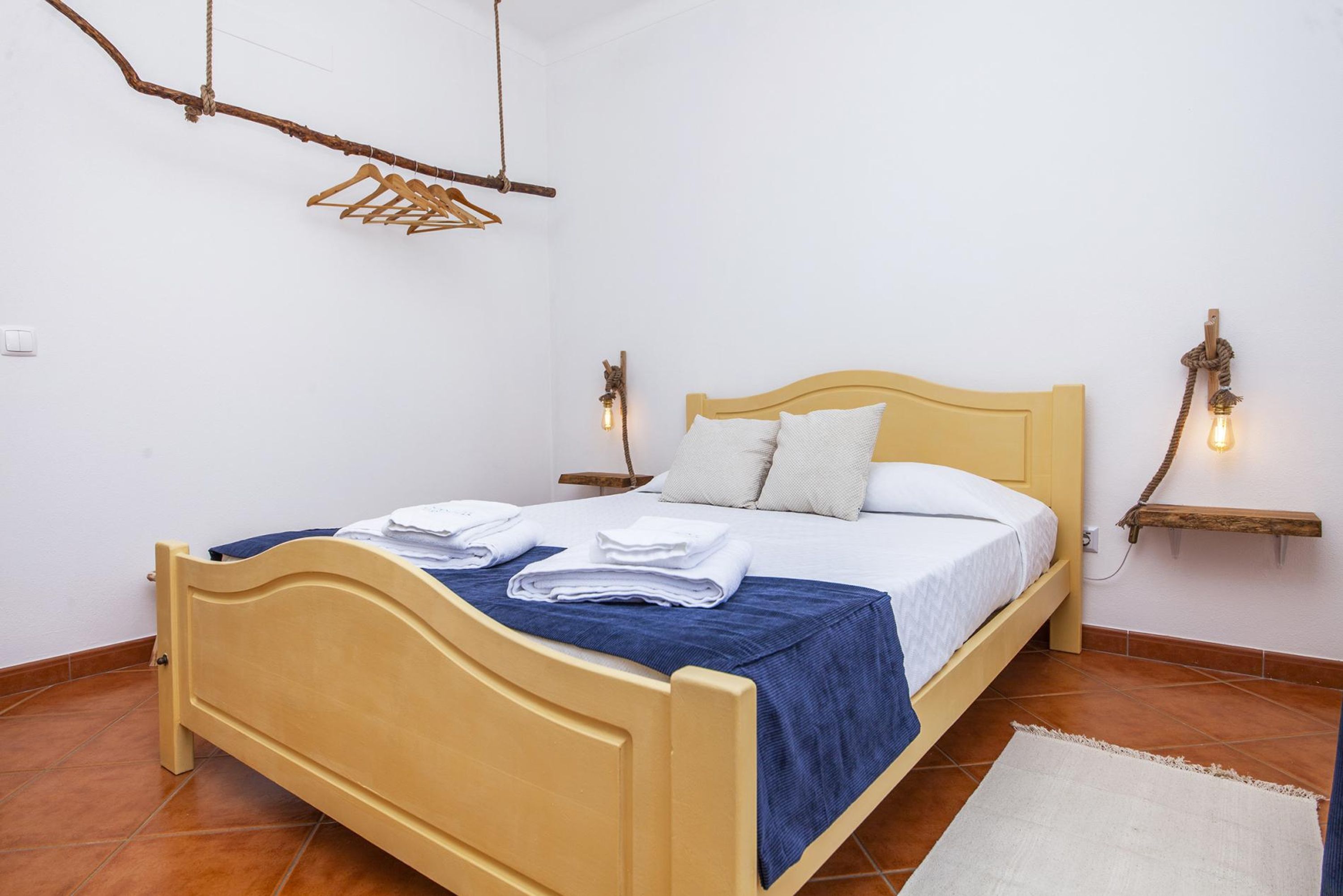 Ein Bett mit hölzernem Kopfteil, einer blauen Bettdecke und weißen Kissen. Zwei Wandleuchten und ein Holzregal mit Seil sind vorhanden.