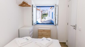 Une petite pièce propre, avec un lit, une commode en bois, une petite fenêtre donnant sur des immeubles et une porte.
