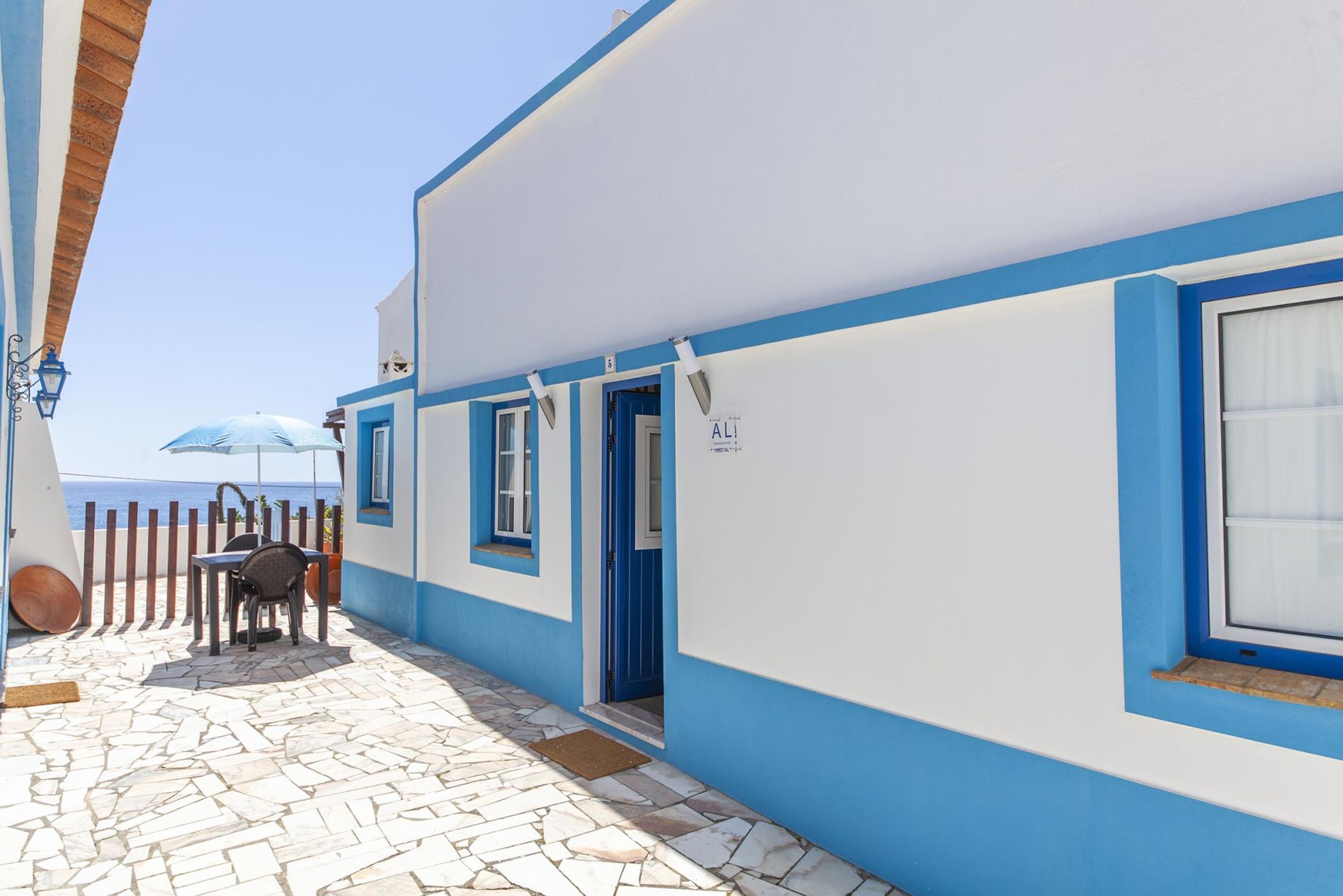Een kustgebouw met een blauw-witte gevel, een patio met stenen bestrating en uitzicht op de zee.