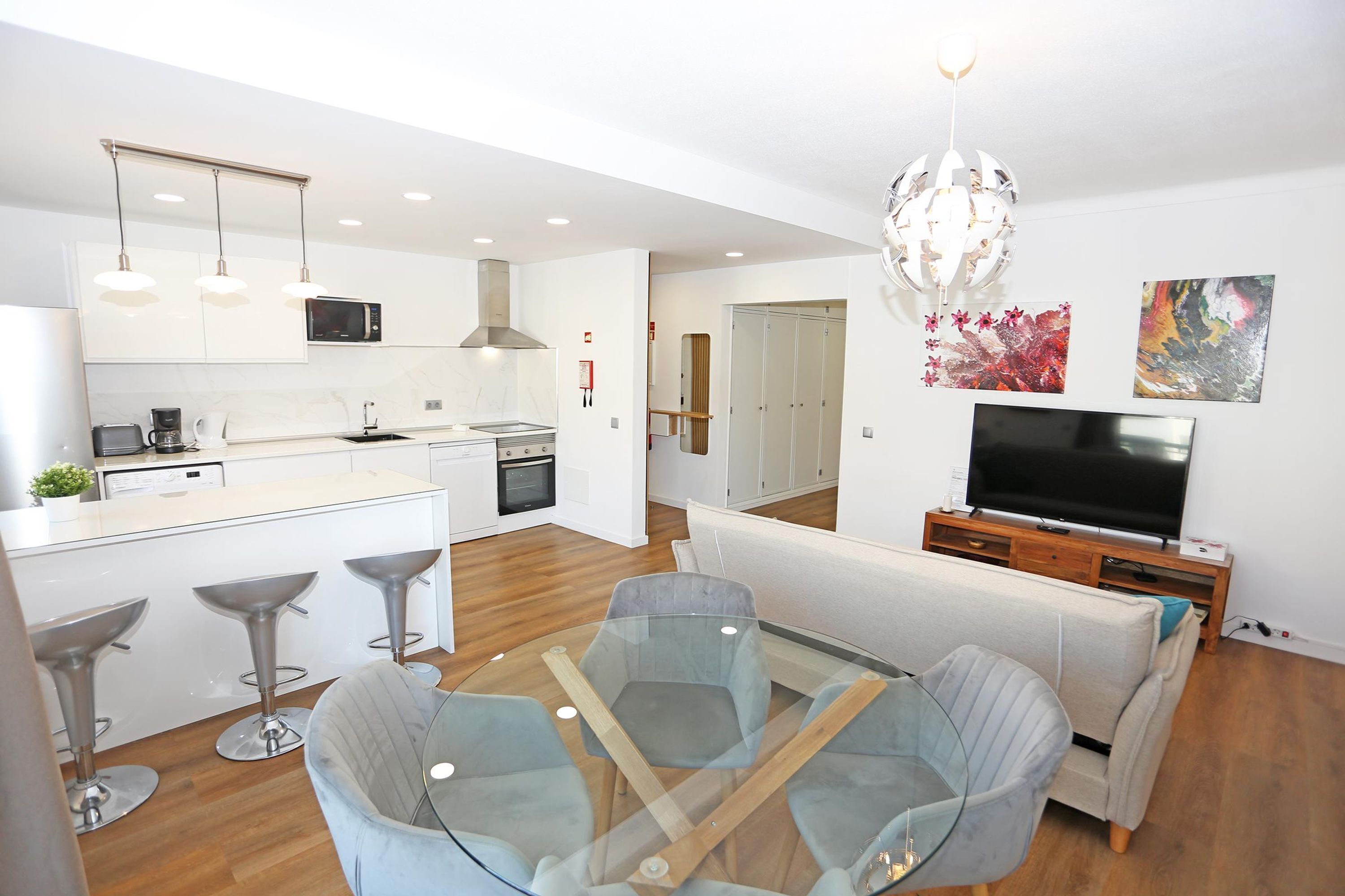 A modern kitchen with white cabinets, a microwave, and a coffee maker. There is a dining area with a glass table and chairs. A TV is mounted on a wooden stand.