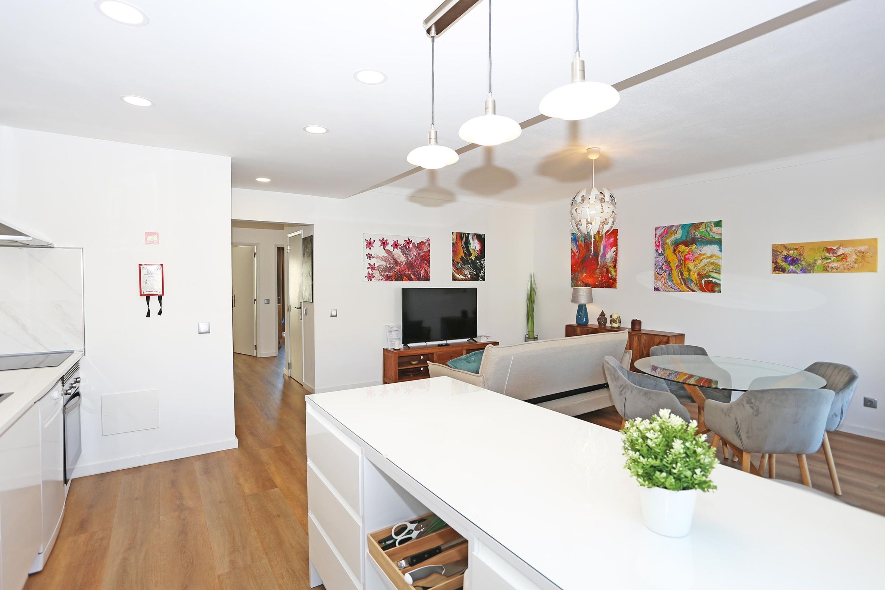 Una cocina moderna con armarios blancos y encimera de madera, un comedor con mesa de vidrio y sillas grises, y una sala de estar con sofá y televisor.