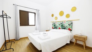 A neatly arranged hotel room with a bed, a bedside table, a nightstand, a window with curtains, and a wall-mounted shelf with decorative items. - A48 - Windmill 1 Bed Apartment in Odiaxere (Lagos)