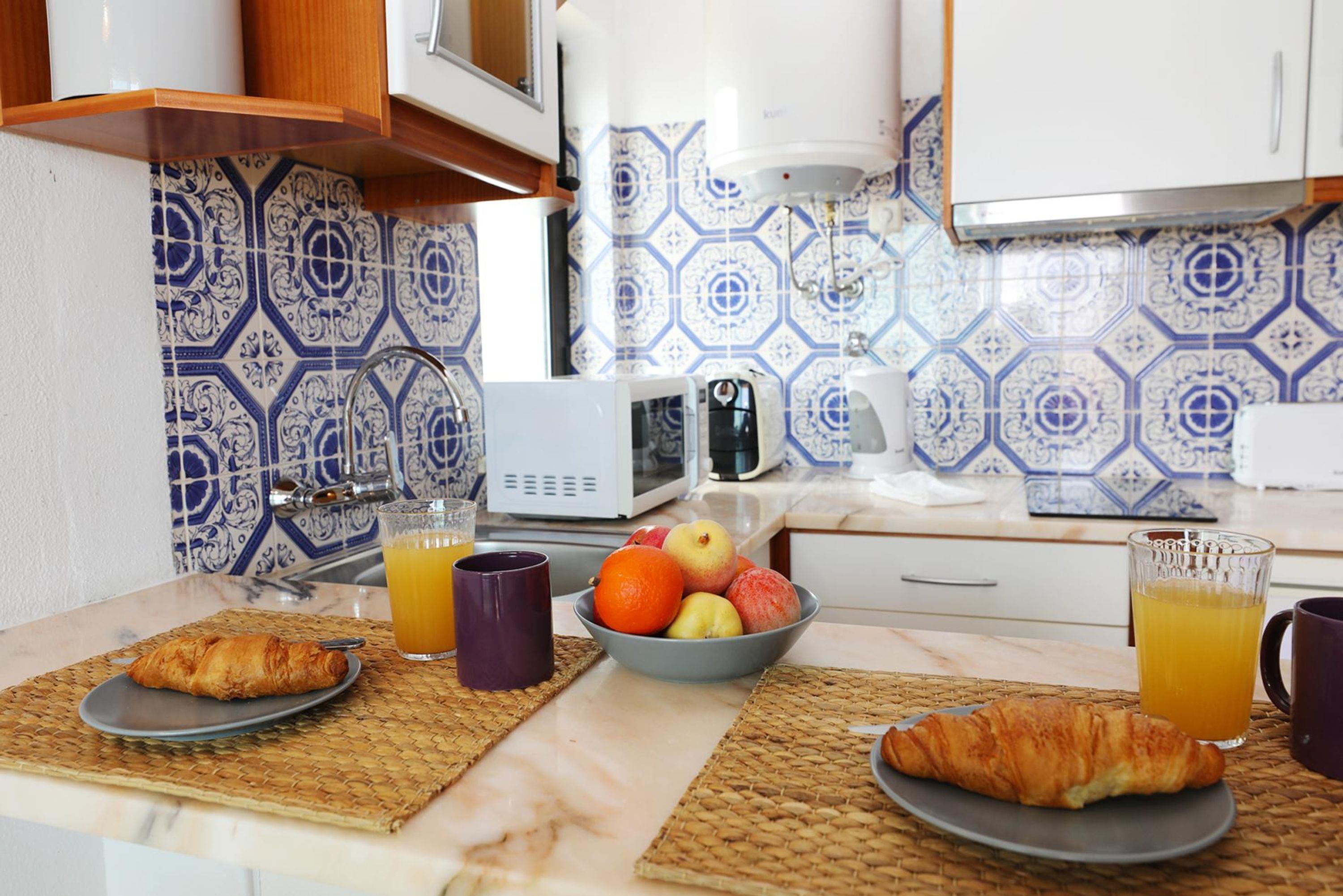 Una cocina con preparación para el desayuno, incluyendo cruasanes, frutas y jugo de naranja.