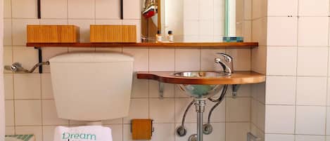 Un baño con inodoro, lavabo y ducha. Hay repisas sobre el inodoro y un espejo sobre el lavabo.