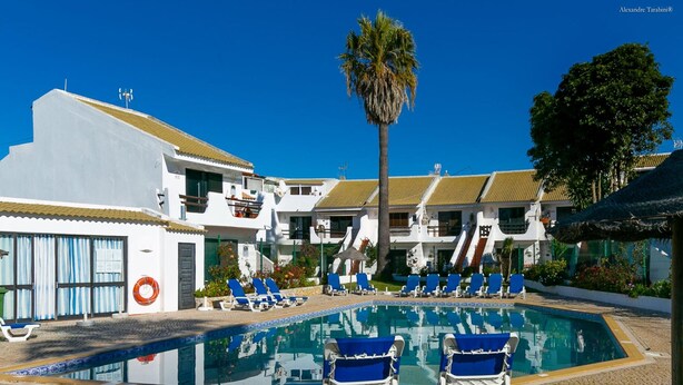 A pool area with blue lounge chairs, a palm tree, and a building with a tiled roof. - B46 - Belavista Torraltinha Flat (Lagos)