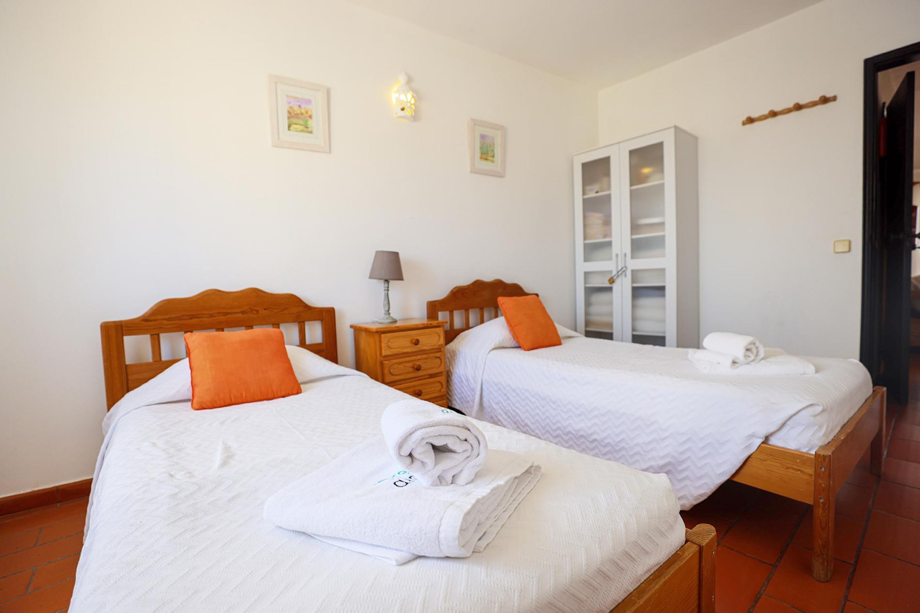 Two single beds with wooden headboards, white bedding, and orange pillows. There is a wooden wardrobe and a nightstand with a lamp.