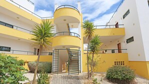 A multi-story building with a yellow facade, a central spiral staircase, and a small garden area with palm trees and shrubs.
