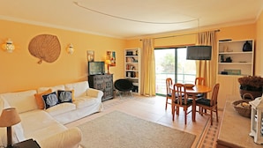 A living room with a sofa, a dining table with chairs, a TV, a bookshelf, and a large window with curtains.