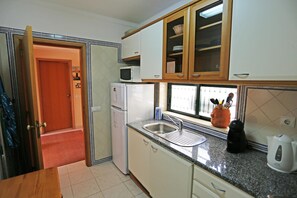 Een kleine keuken met witte kastjes, een ingebouwde magnetron, een koelkast, een spoelbak en een raam met uitzicht.