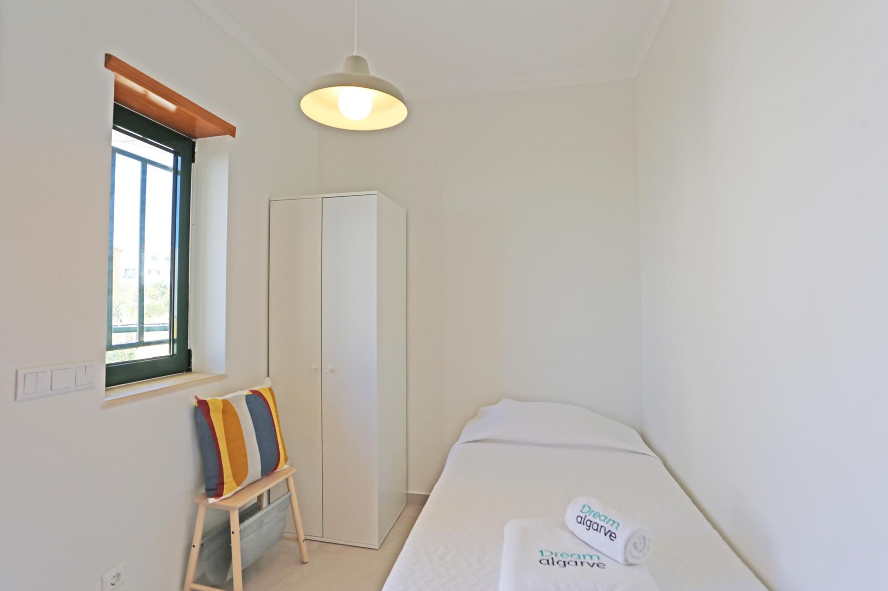 A small, clean room with a bed, a wardrobe, a window, and a small table with a pillow.