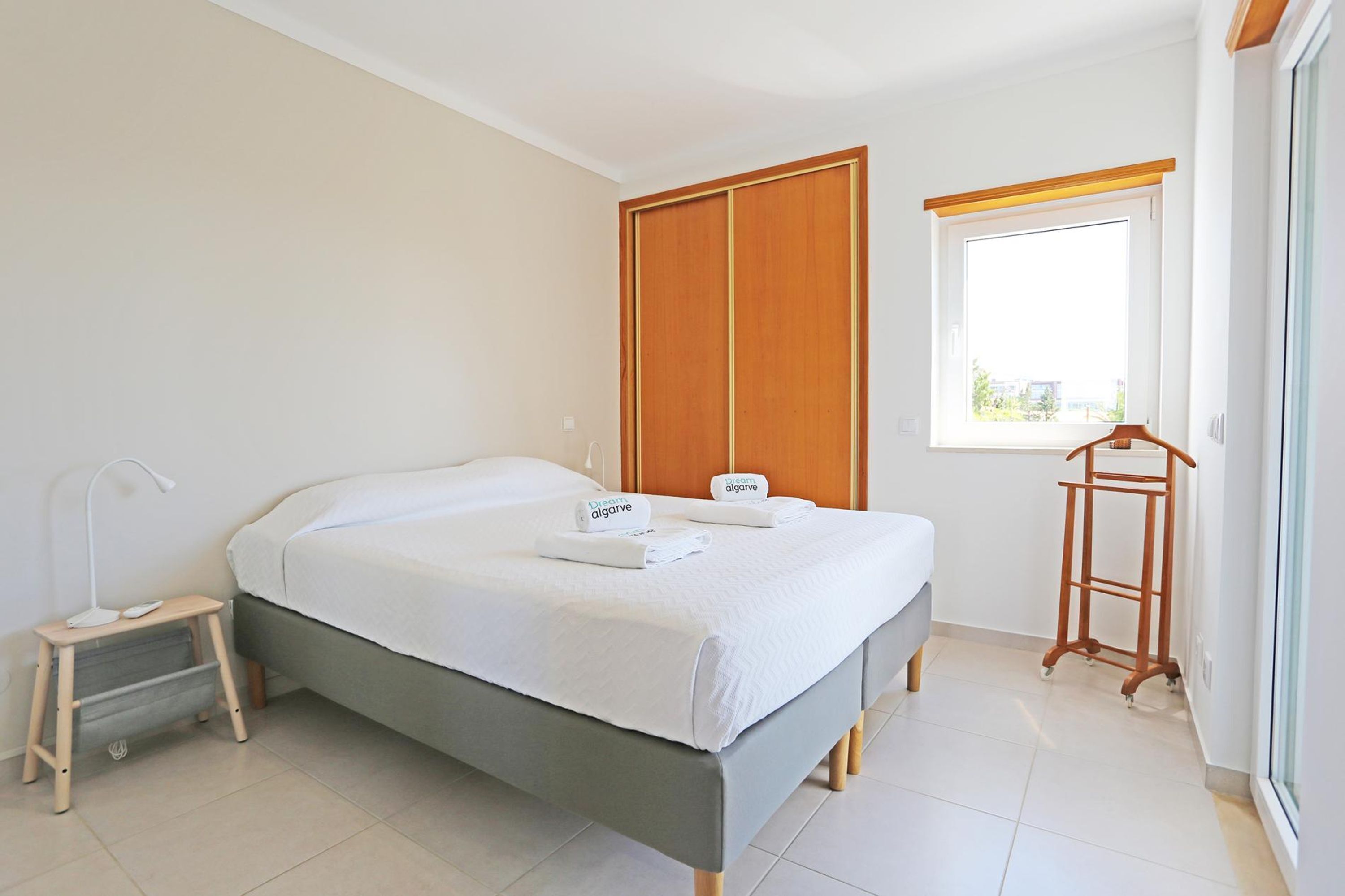 A neatly made bed with a grey headboard, a bedside table with a lamp, and a wooden wardrobe.