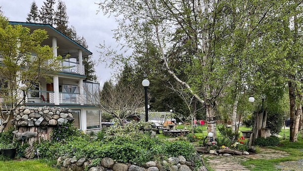 Un sendero de piedra conduce a un muro de piedra rústico con jardín, una terraza de madera y un edificio con balcón.