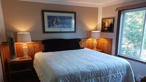 Un dormitorio con una cama grande, dos lámparas de noche, un cuadro enmarcado sobre la cama y una ventana con vistas a árboles.