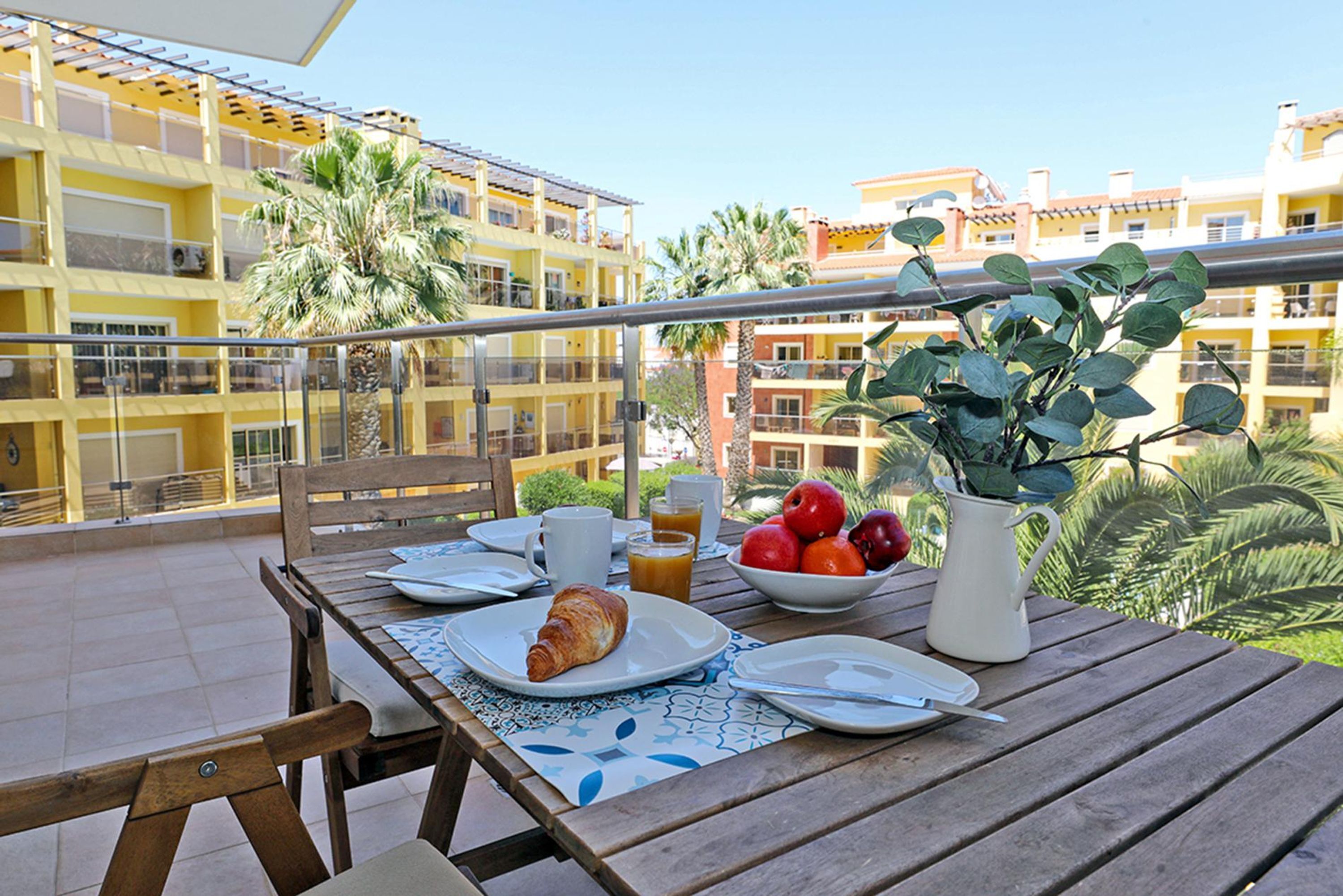 A balcony with a wooden table set for breakfast, including croissants, fruit, and a pitcher.