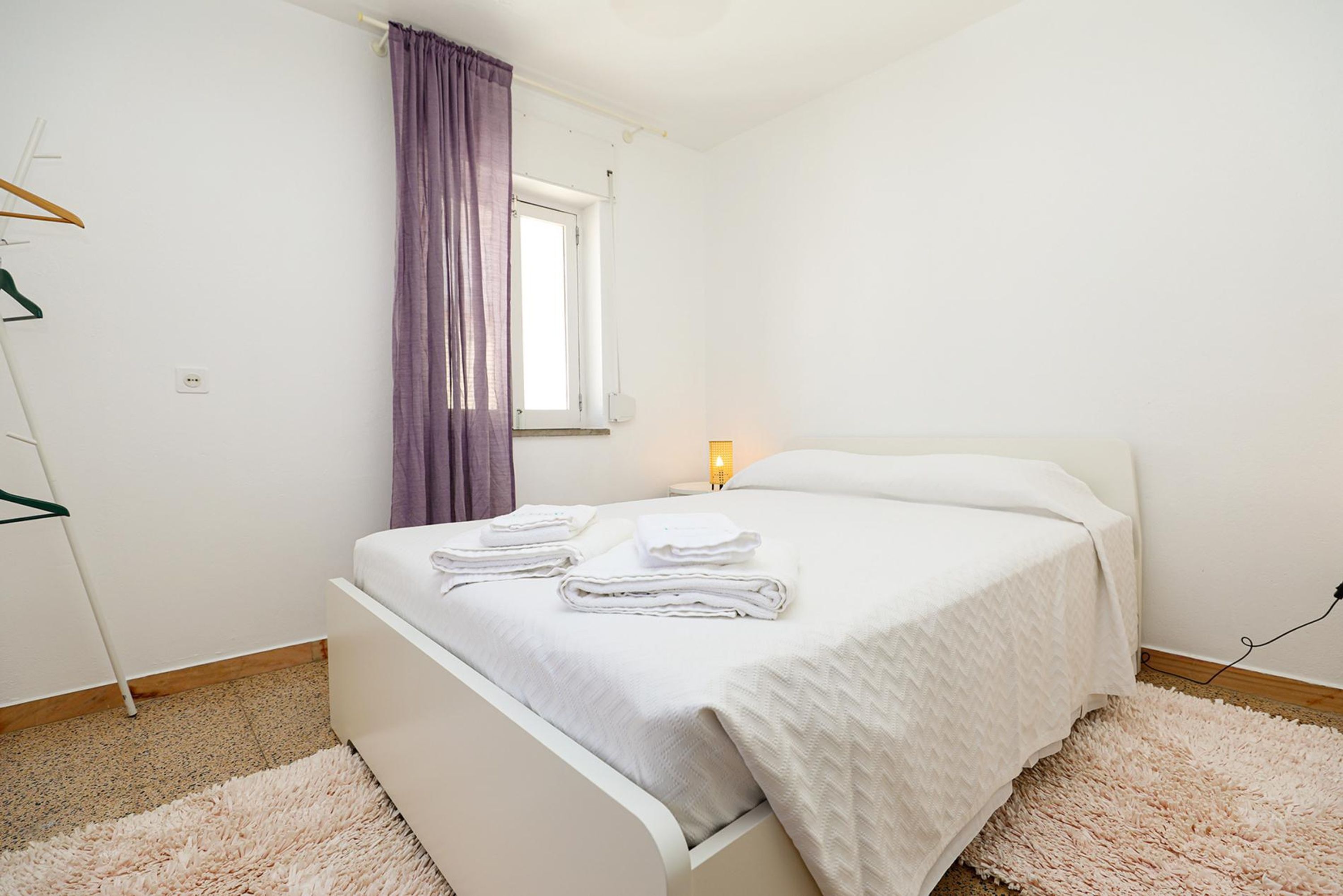 A bedroom with a bed, a window with curtains, a bedside lamp, and a white wardrobe.