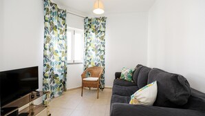 Una sala de estar con televisor de pantalla plana, sofá gris oscuro, sillón de mimbre y cortinas con estampado tropical.
