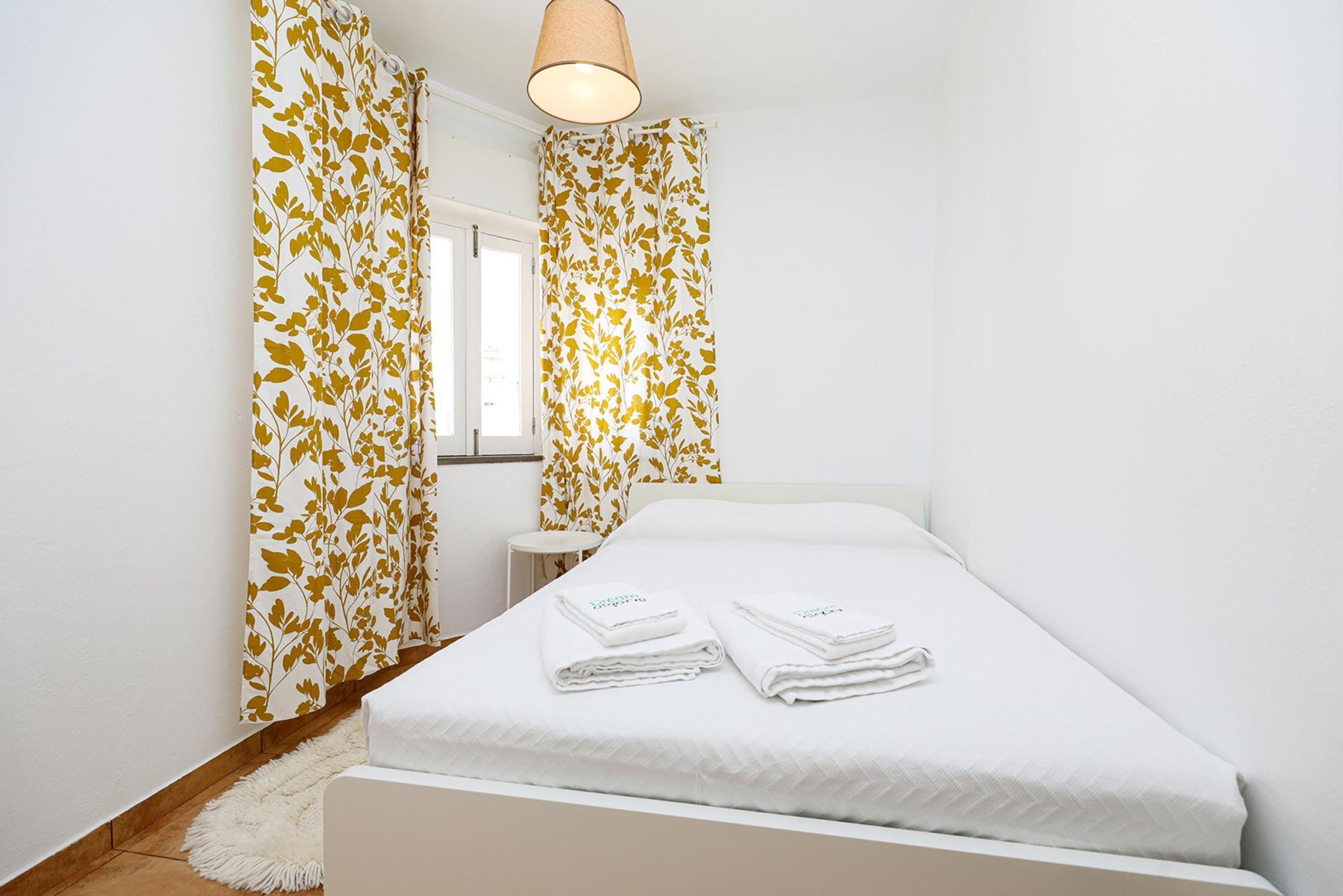 A hotel room with a bed, white curtains with a leaf pattern, a small bedside table, and a lamp.