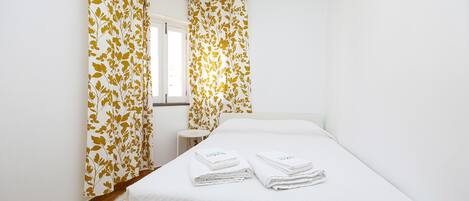 A hotel room with a bed, white curtains with a leaf pattern, a small bedside table, and a lamp.
