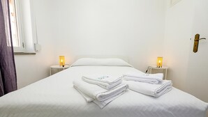 Una cama bien hecha con toallas blancas dobladas, dos mesitas de noche con lámparas y un ventanal con cortinas.