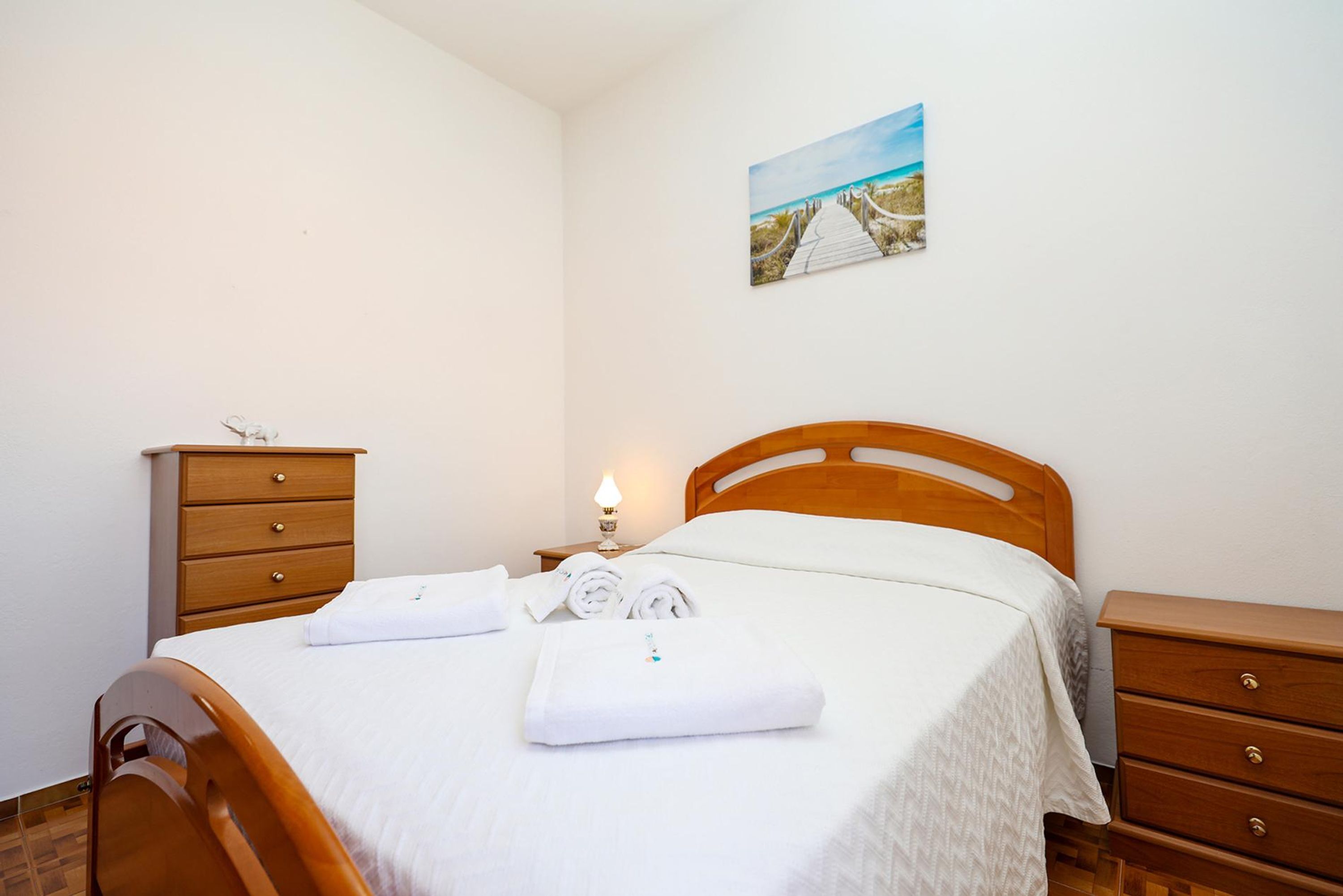 A neatly made bed with white linens and towels, a wooden headboard, a nightstand with a lamp, and a small wooden dresser.