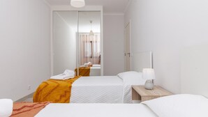 A hotel room with two beds, a mirror, a bedside table with a lamp, and a view into the adjacent room.