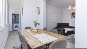 A modern living room with a dining area, a black sofa, a clock on the wall, and a washing machine in the background.
