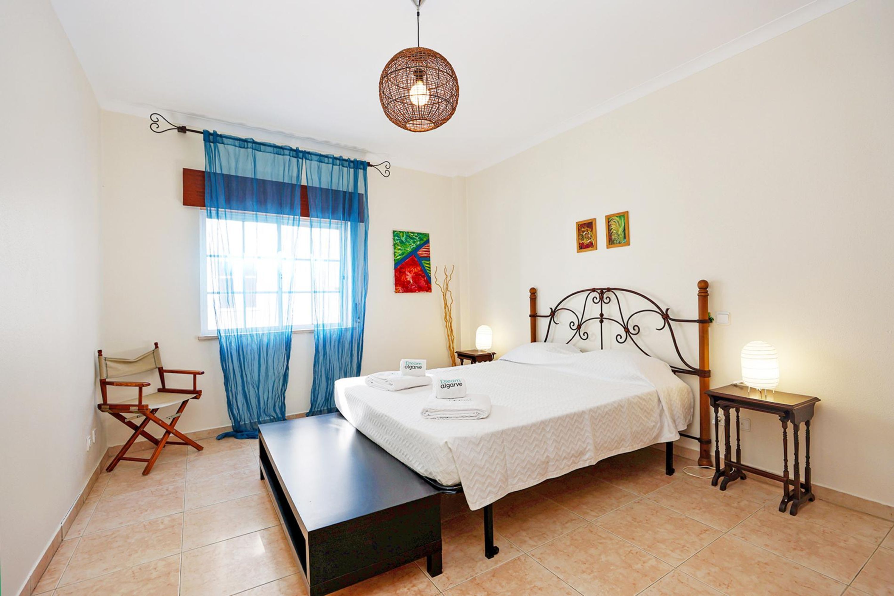 A bedroom with a bed, a bench, a director's chair, a bedside table with a lamp, and a window with curtains.