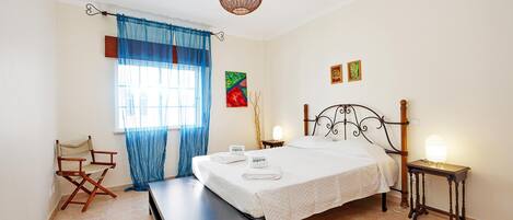 A bedroom with a bed, a bench, a director's chair, a bedside table with a lamp, and a window with curtains.