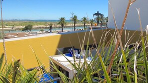 A balcony with a view of the sea, palm trees, and a beach area with umbrellas and a gazebo. - C25 - Town Center Apartment in Lagos (Lagos)