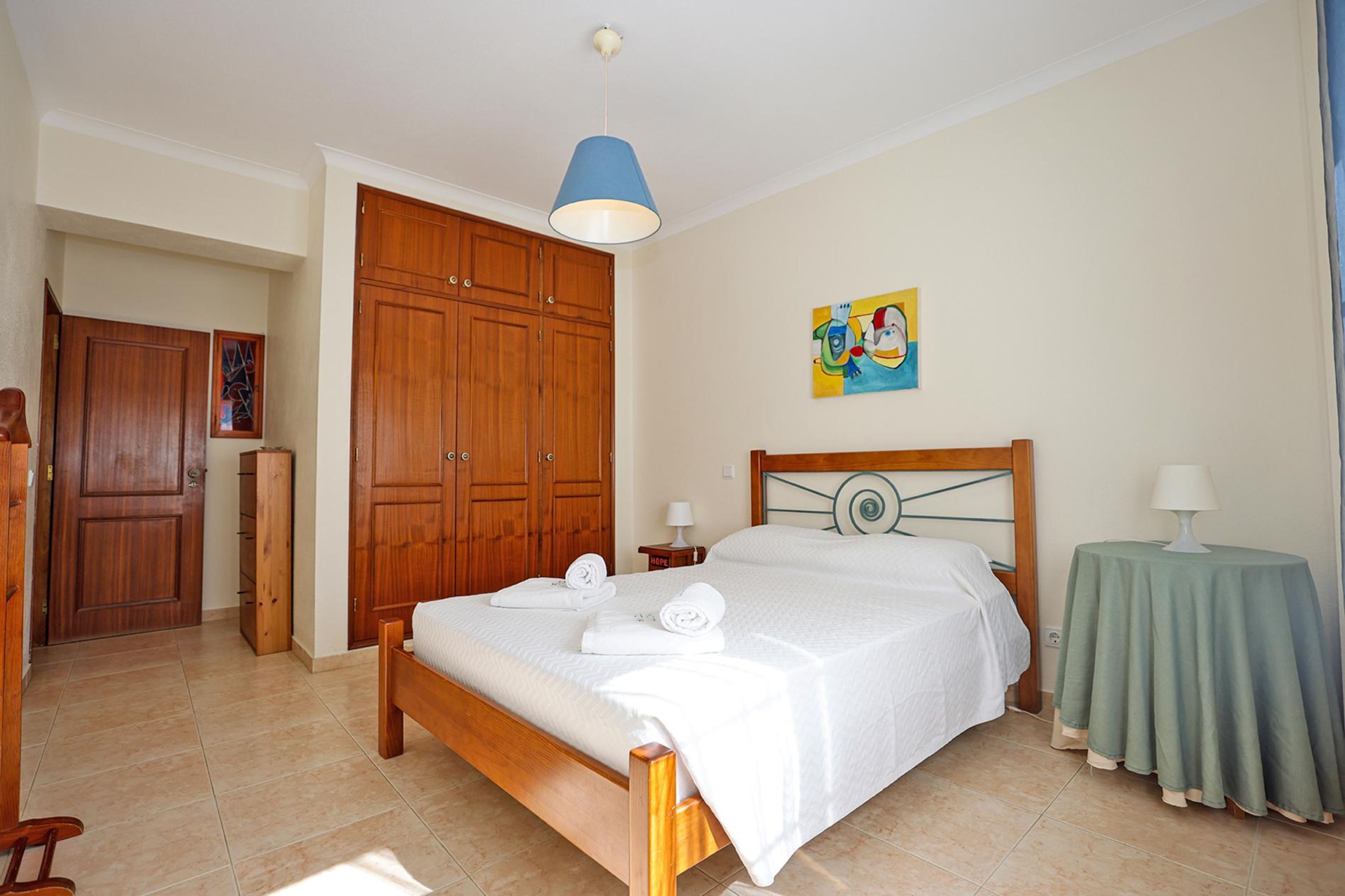 A bedroom with a wooden bed, a blue hanging light, a green side table, and a painting on the wall.