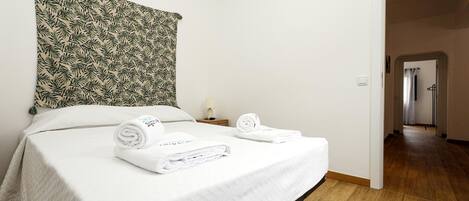 Une chambre d’hôtel avec un lit, deux serviettes enroulées, une table de chevet avec une lampe et une tapisserie à motifs.