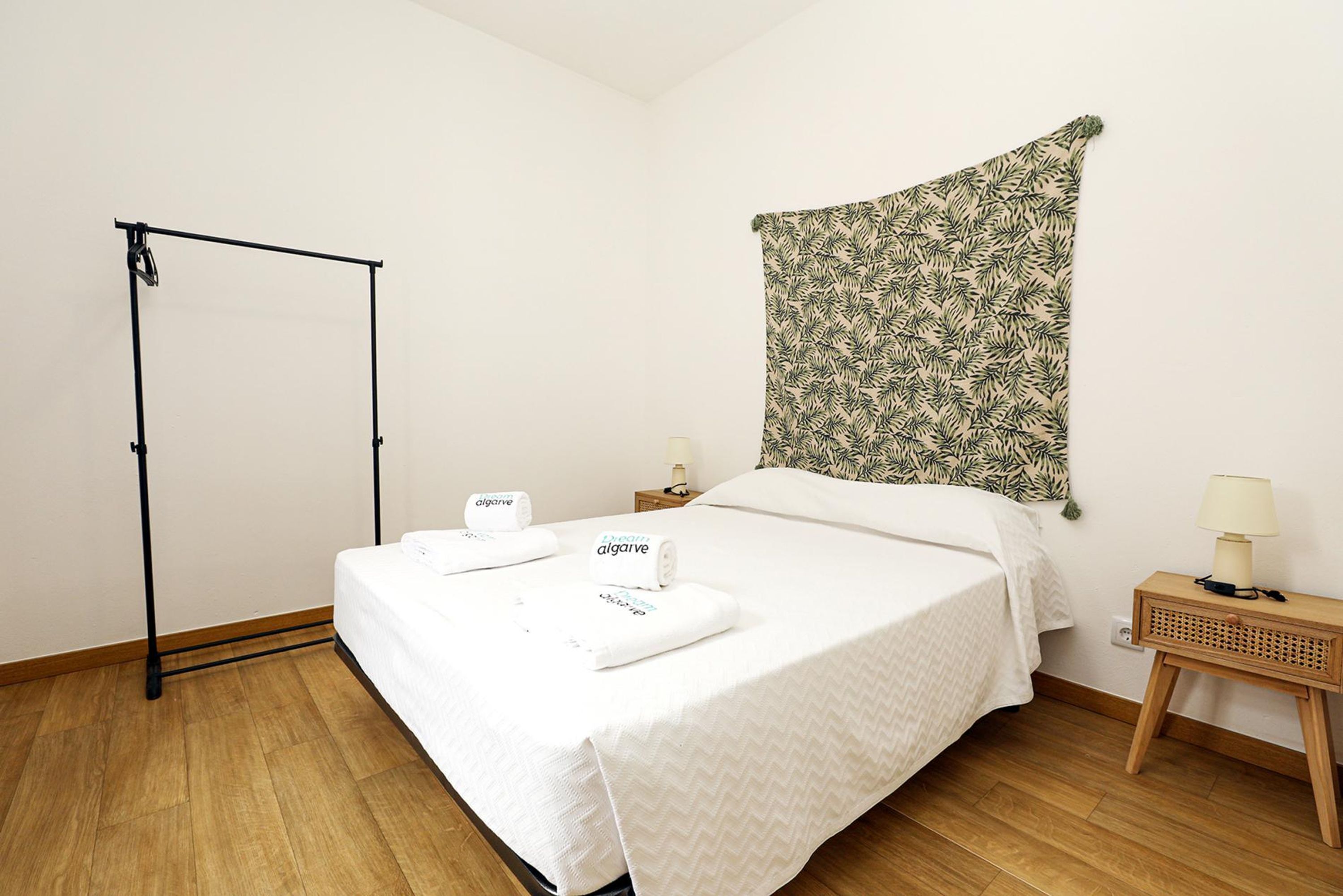 A neatly made bed with white linens and two towels, a wooden floor, a wall-mounted tapestry, a small bedside table with a lamp, and a black metal stand.