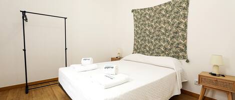A neatly made bed with white linens and two towels, a wooden floor, a wall-mounted tapestry, a small bedside table with a lamp, and a black metal stand.