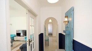 A hallway with a white door, a blue door, a bench, a television in a living room, and a chandelier. - C13 - Ameijeira Holiday Apartment in Lagos (Lagos)