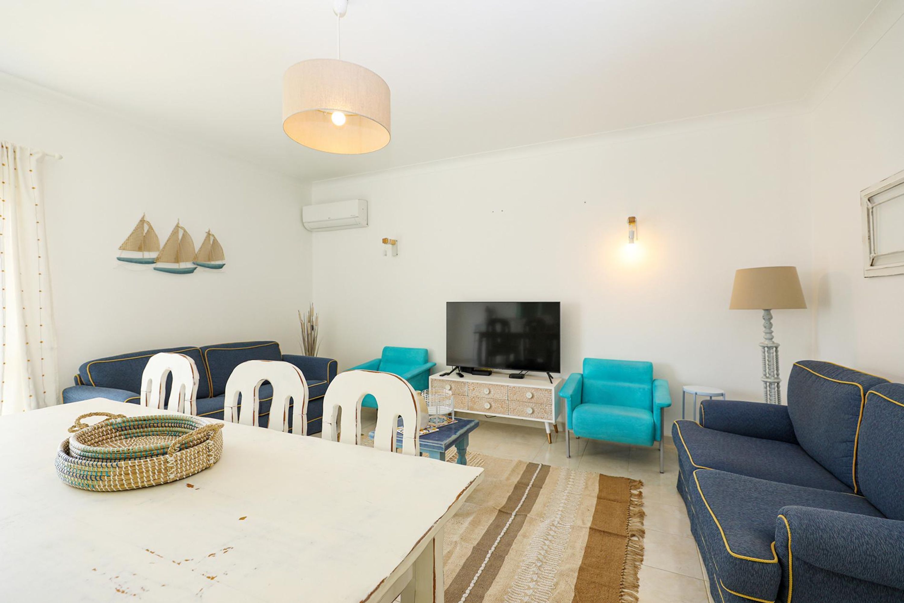 Una sala de estar con una mesa blanca, sofás azules, un televisor y objetos decorativos.