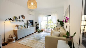 A modern living room with a sofa, a coffee table, and a TV. There is a large window with curtains, a hanging light fixture, and several potted plants. - B95 - Casa Melo Apartment in Praia da Luz (Lagos)