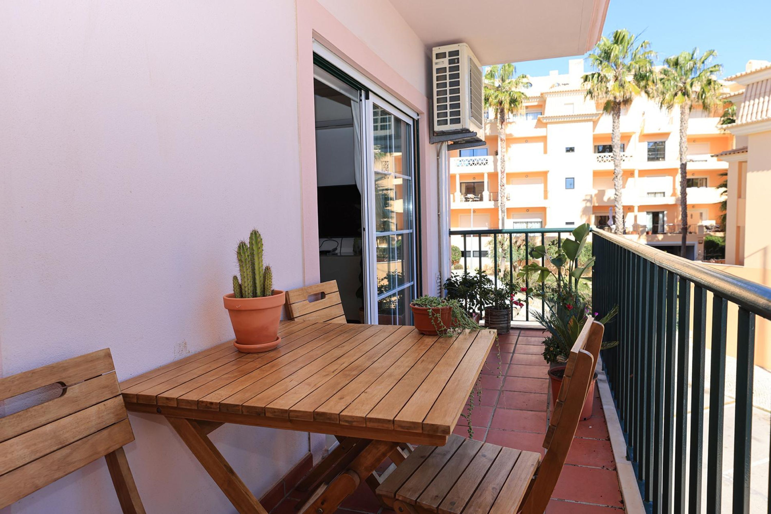 A wooden table with chairs and potted plants on a balcony with a view of palm trees and a building in the background.