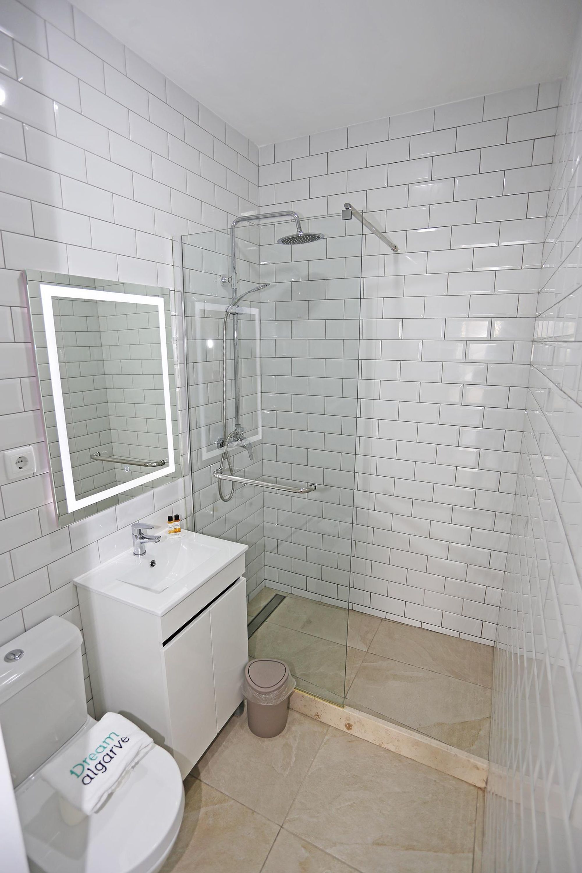 A bathroom with white tiled walls, a glass-enclosed shower, a white sink, and a toilet with a 'Dream Alugarve' sign.