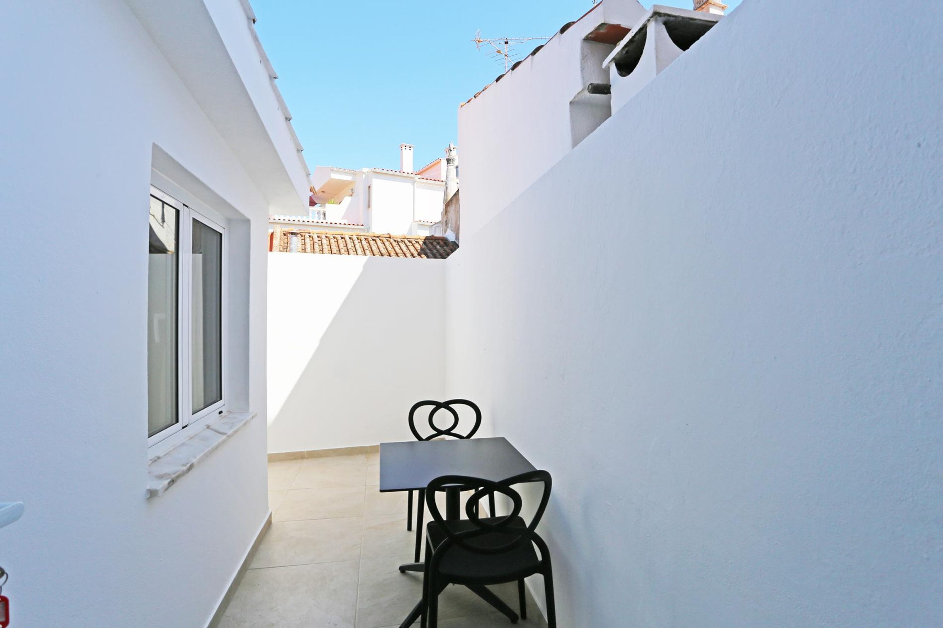 A narrow outdoor corridor with a white wall, a window, and a table with two chairs.