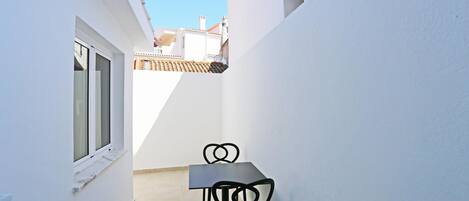 A narrow outdoor corridor with a white wall, a window, and a table with two chairs.