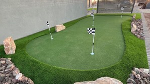 A well-maintained artificial green lawn with three checkered flags, surrounded by rocks and a building with a gray facade. - 2-bedroom Home-casita , private backyard with grill, putting green,cornhole (Buckeye)