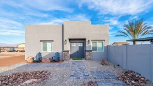 A modern two-story house with a grey facade, a front door with a green mat, and a small garden area with rocks and a paved walkway. - 2-bedroom Home-casita , private backyard with grill, putting green,cornhole (Buckeye)