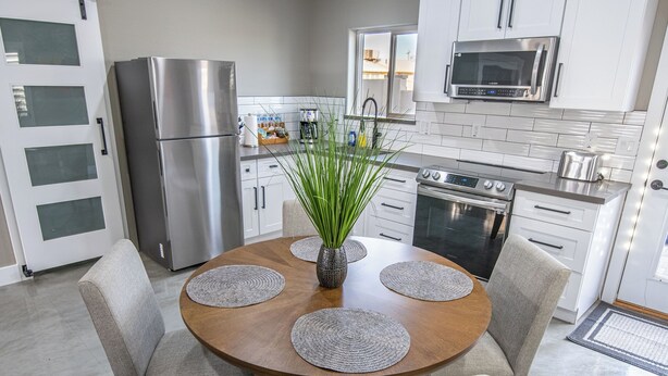 A modern kitchen with white cabinets, stainless steel appliances, and a round dining table with placemats. - 2-bedroom Home-casita , private backyard with grill, putting green,cornhole (Buckeye)