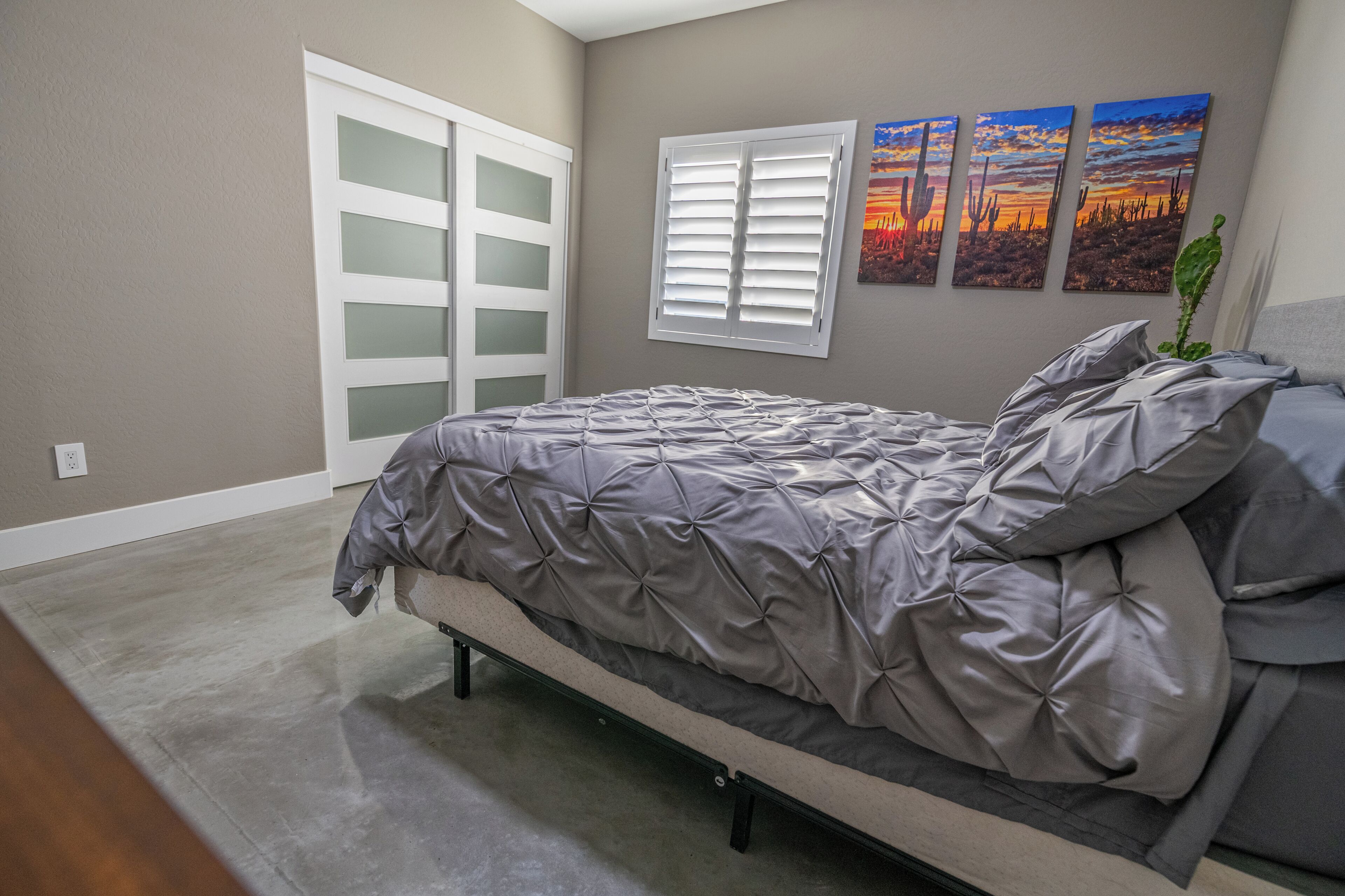 A bedroom with a bed, a closet with frosted glass doors, a window with blinds, and three desert landscape paintings on the wall.